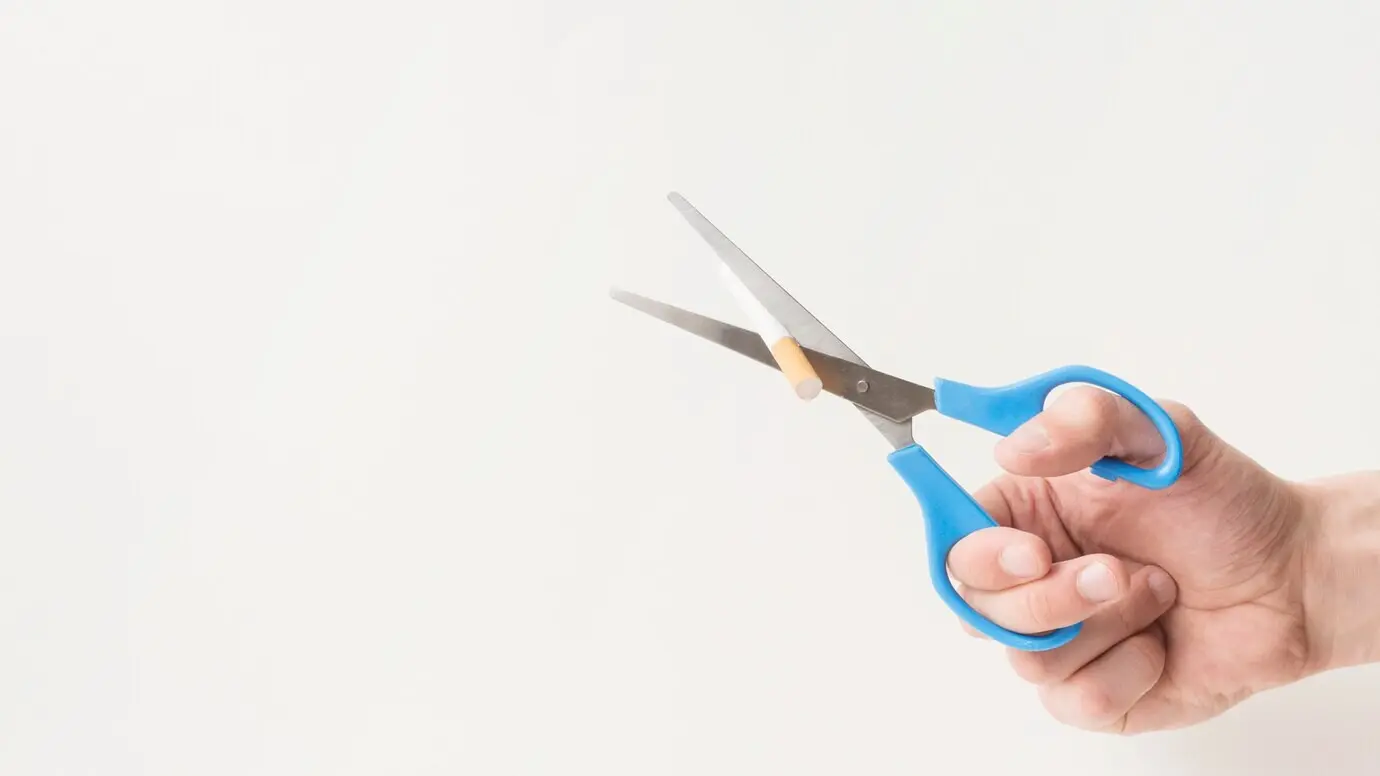 A man's hand uses scissors to cut a cigarette on a white surface.